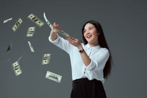 soldi sorridente ragazza asiatica gettando banconote in dollari isolate su grigio