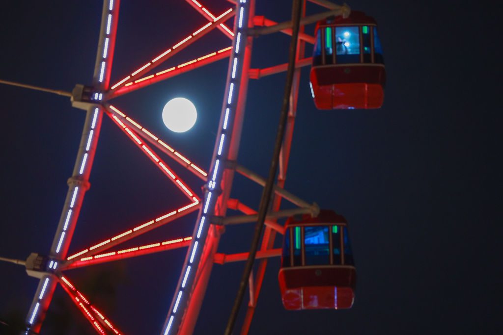 Primo piano di una ruota panoramica rossa con le luci accese e la luna che splende dietro di essa.