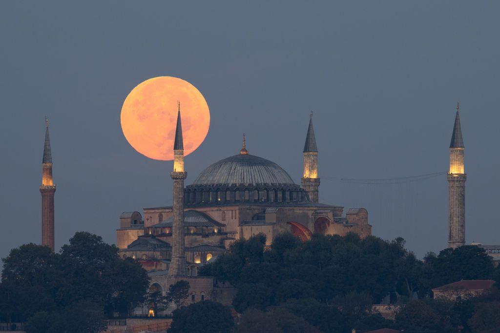 La luna piena bassa nel cielo risplende di un colore leggermente rosa-arancio sopra una grande moschea.
