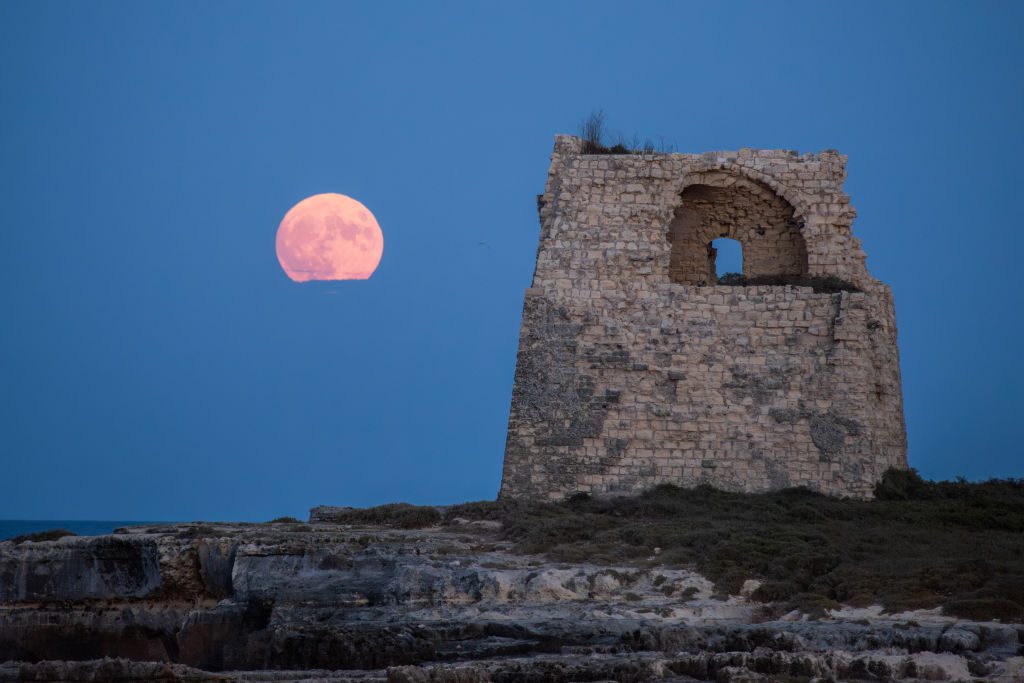 La gigantesca luna blu a sinistra brilla di rosa accanto a una struttura a torre in pietra con una piccola apertura in alto.
