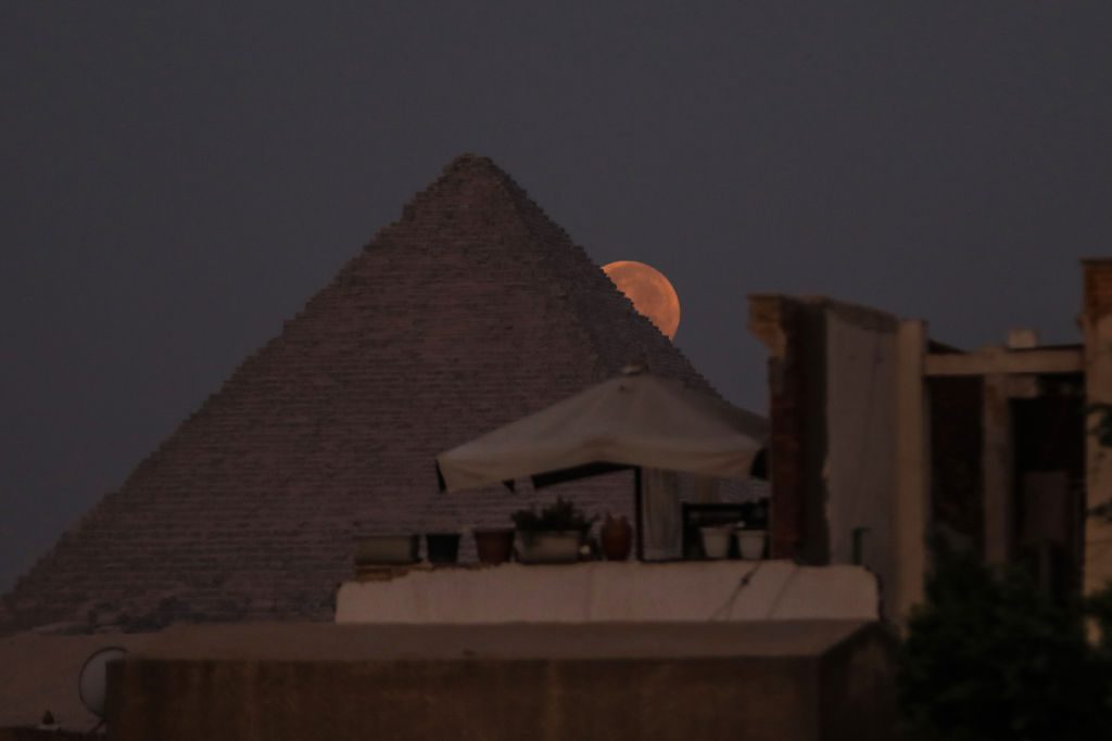 Una luna piena rosso/rosa arancione appare dal lato della Grande Piramide.