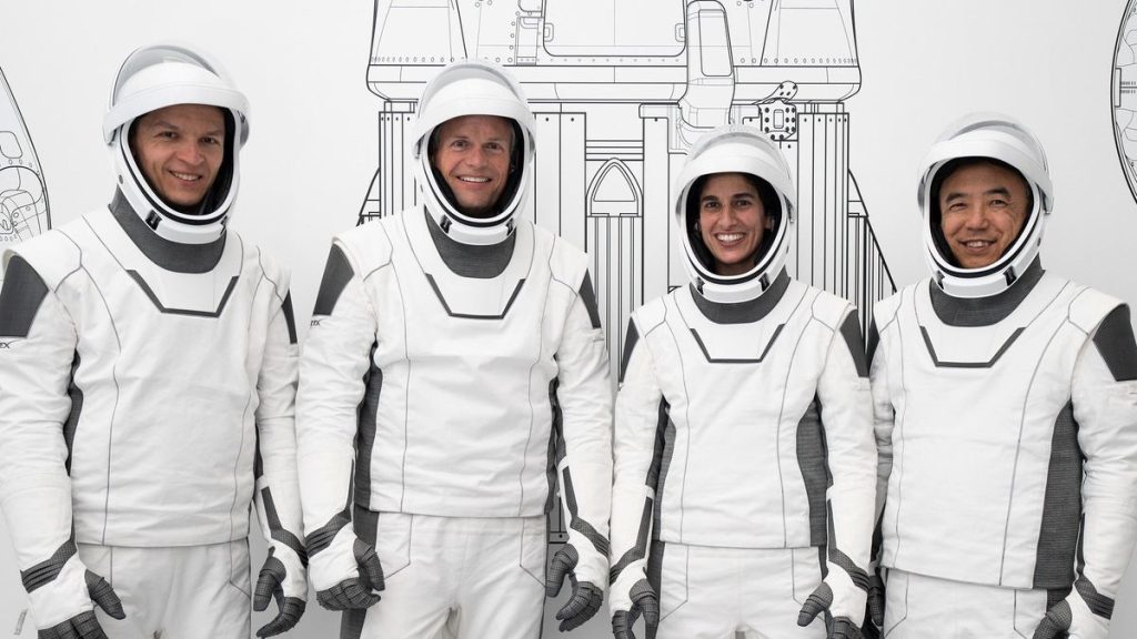 four astronauts standing in a row in white spacesuits and smiling. a line drawing of a rocket is partially visible behind them