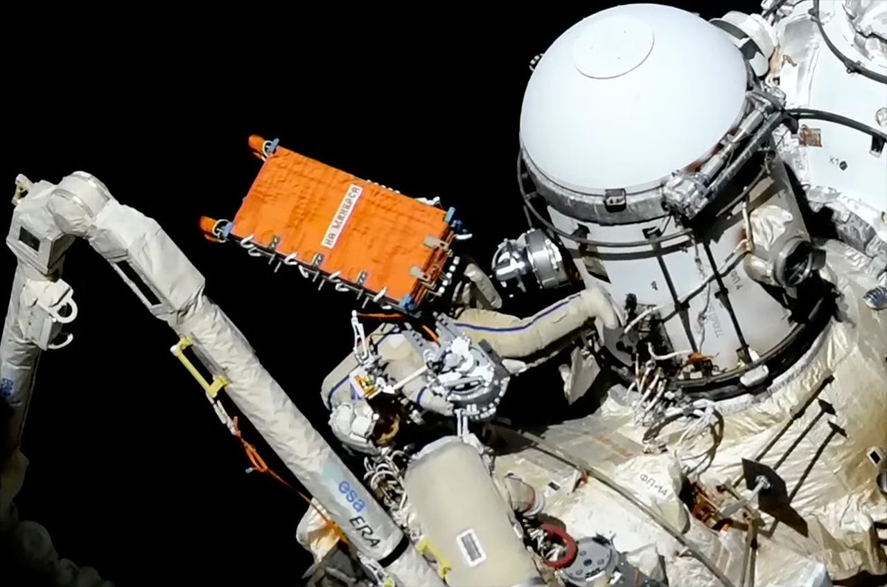Un astronauta che indossa una tuta spaziale bianca installa un sistema di comunicazione radar arancione sulla Stazione Spaziale Internazionale.
