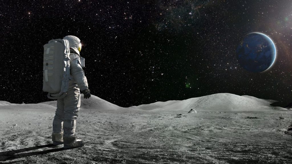 A lone astronaut standing facing away from the camera dressed in full space suit with backpack, stands still looking towards a distant planet Earth.