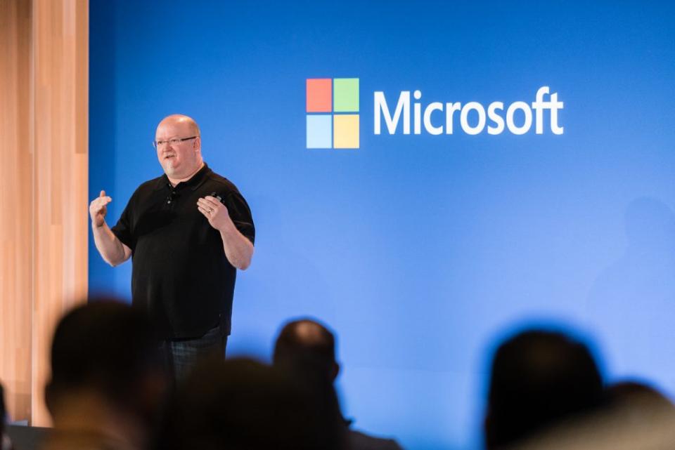 Il CTO di Microsoft Kevin Scott tiene una presentazione sul palco davanti a un muro blu con il logo Microsoft.  Le teste del pubblico sono sfocate in primo piano.